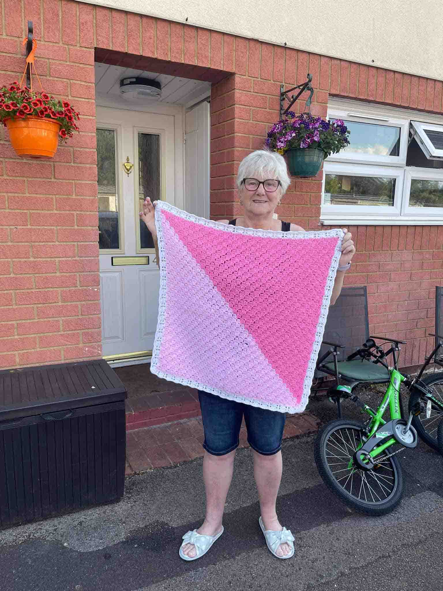 Person called Enid stands outside entrance to a residential building, holding up to camera a pink baby blanket she has knitted for clients of The Dash Charity.