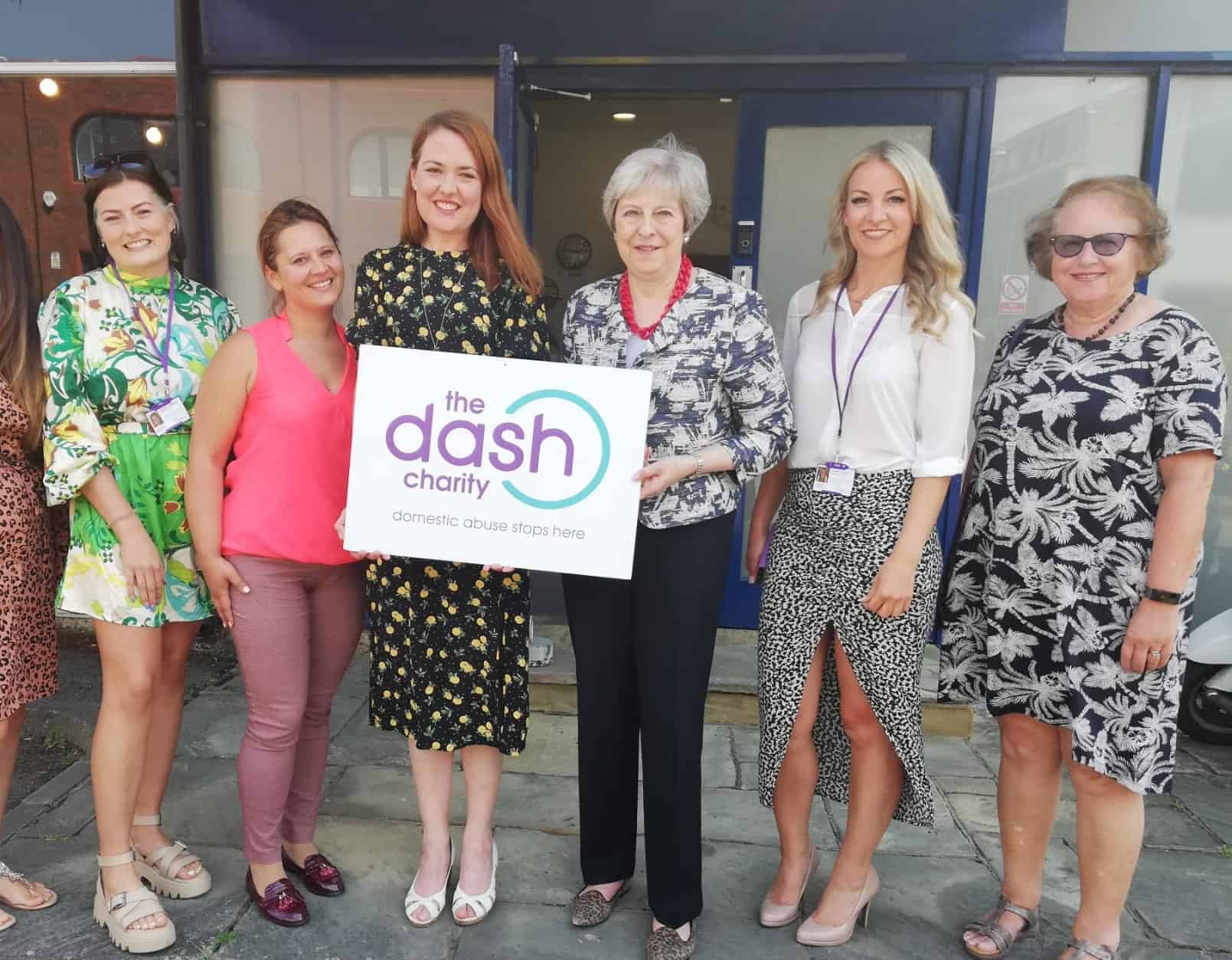 Six women stand facing the camera, including CEO Nicola Miller and Theresa Map MP in the centre holding The Dash Charity logo on a small sign.