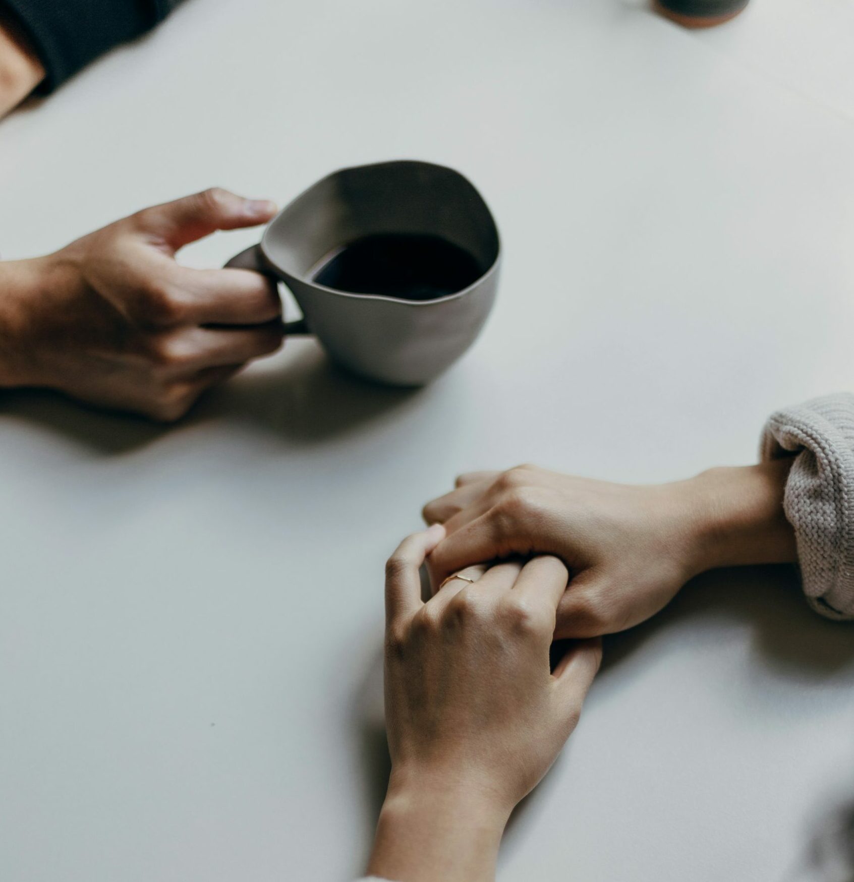 Two people sit at a table, one holding a mug of coffee, while the other person's hands are clasped together nearby.