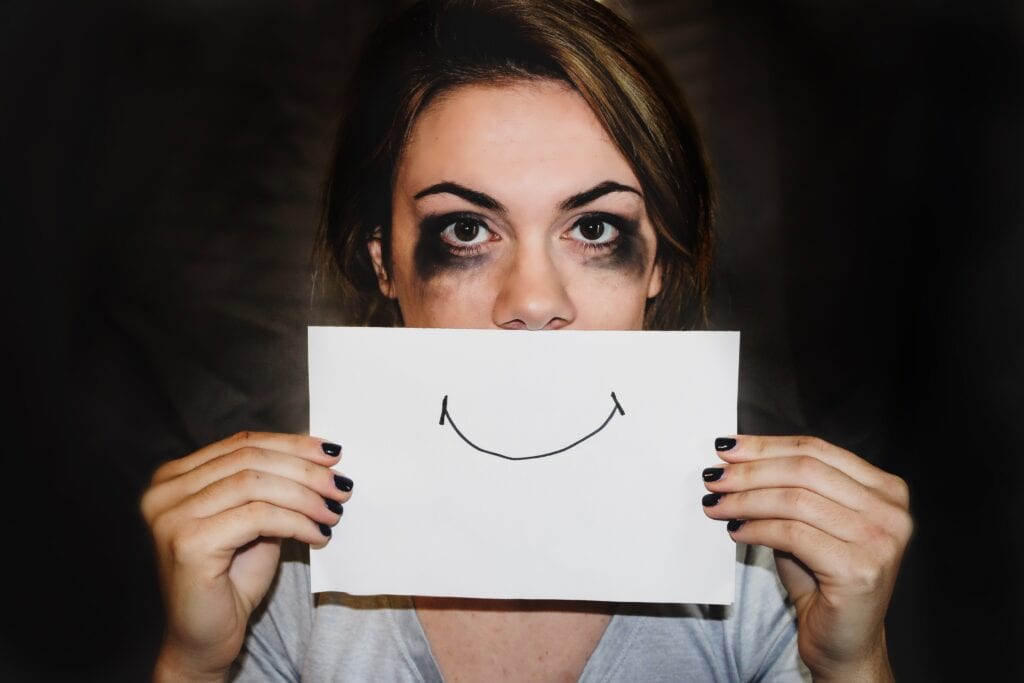 Female presenting person holds white piece of A4 paper in front of her mouth with a hand drawn smile on it