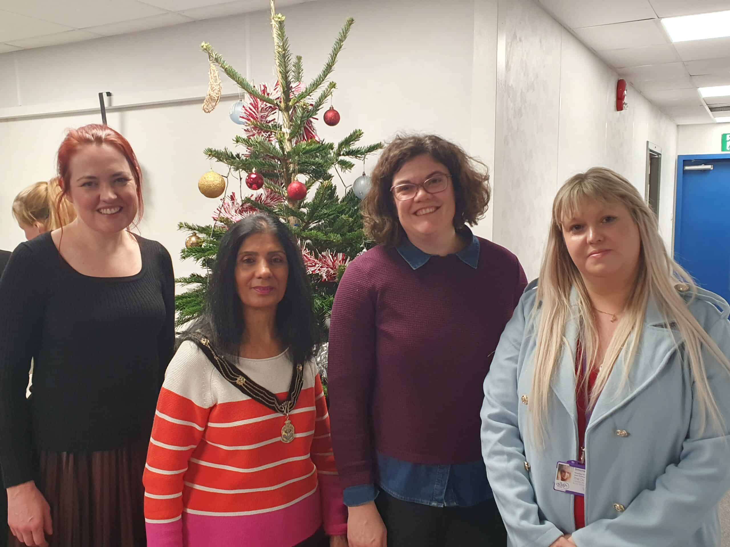 Four women stand in front of a decorated Christmas tree at a festive event. From left to right: Nicola Miller, CEO of The Dash Charity, wearing a black top and smiling; Mandy Brar, Deputy Mayor of the Royal Borough of Windsor and Maidenhead, in a red and white striped jumper with her ceremonial chain; Cllr Amy Tisi, in a burgundy jumper and glasses; and Simone Butterworth, Housing IDVA at The Dash Charity, wearing a light blue coat and badge. The hallway setting features festive decorations and a blue door in the background