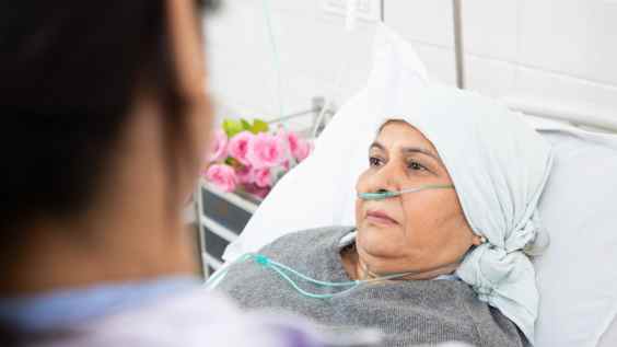 A woman lying in a hospital bed, wearing a light-coloured headscarf and an oxygen tube. She appears calm, gazing towards a person in the foreground, possibly a healthcare professional. The background includes a vase of pink flowers on a bedside table, adding a comforting touch to the clinical setting