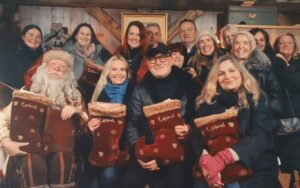 A group photo featuring smiling individuals gathered around Santa Claus in a cosy, festive wooden setting. Santa is seated at the centre, holding a 'Good List' book, while the group holds embroidered red stockings with the word 'Lapland' on them. The scene includes warm decorations, and the text 'Lapland 2024' is displayed at the bottom of the image in golden lettering