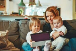 A mother with short red hair sits on a grey sofa with her two young children, a toddler and a baby, in a cosy home setting. The toddler, dressed in white, holds a tablet, while the baby, also in white, looks at the screen with curiosity. The mother smiles as she watches with them, creating a warm and engaged family moment.