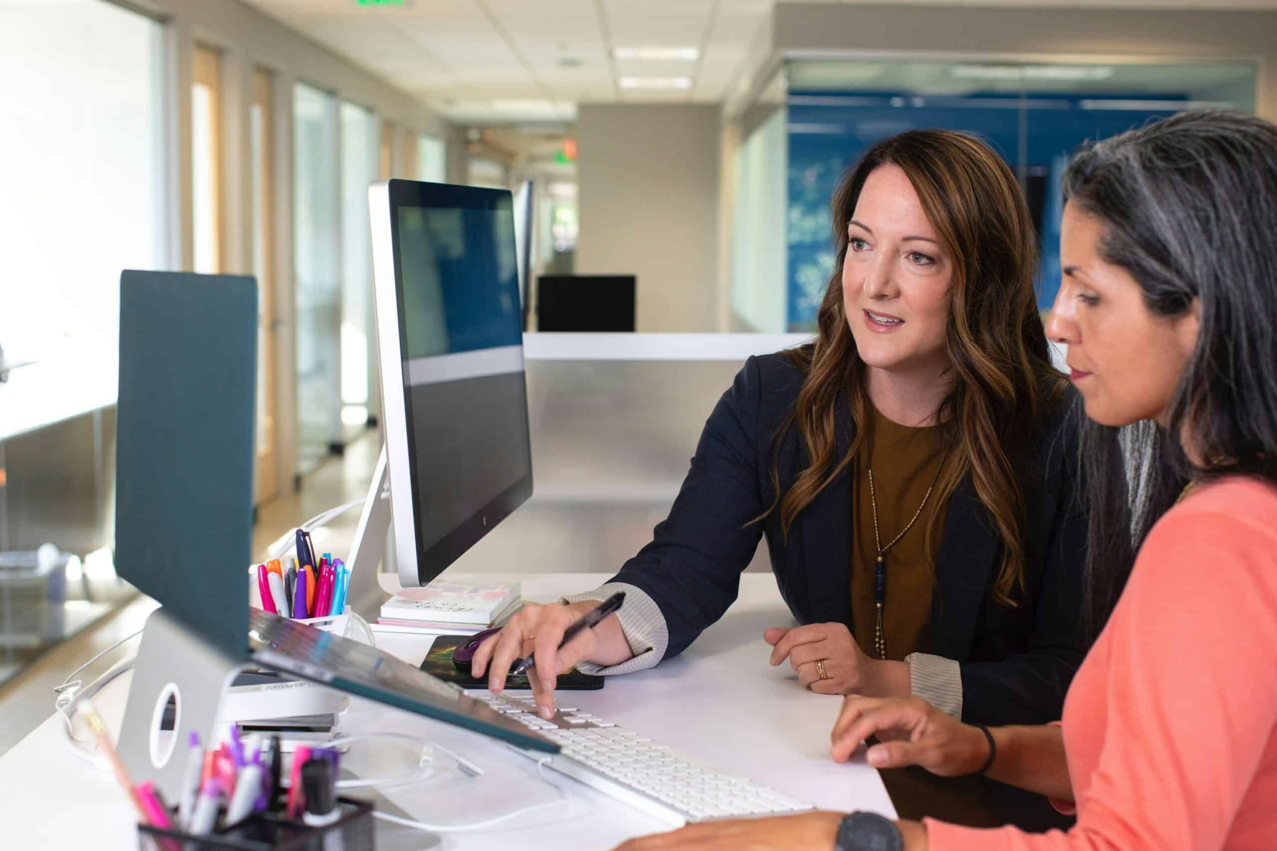 Two professional women in an office setting, sitting at a desk with a computer. One woman, wearing a dark blazer, is pointing at the screen while explaining something to the other, who is dressed in a coral-coloured top. The workspace is modern and well-lit, with office supplies neatly arranged on the desk