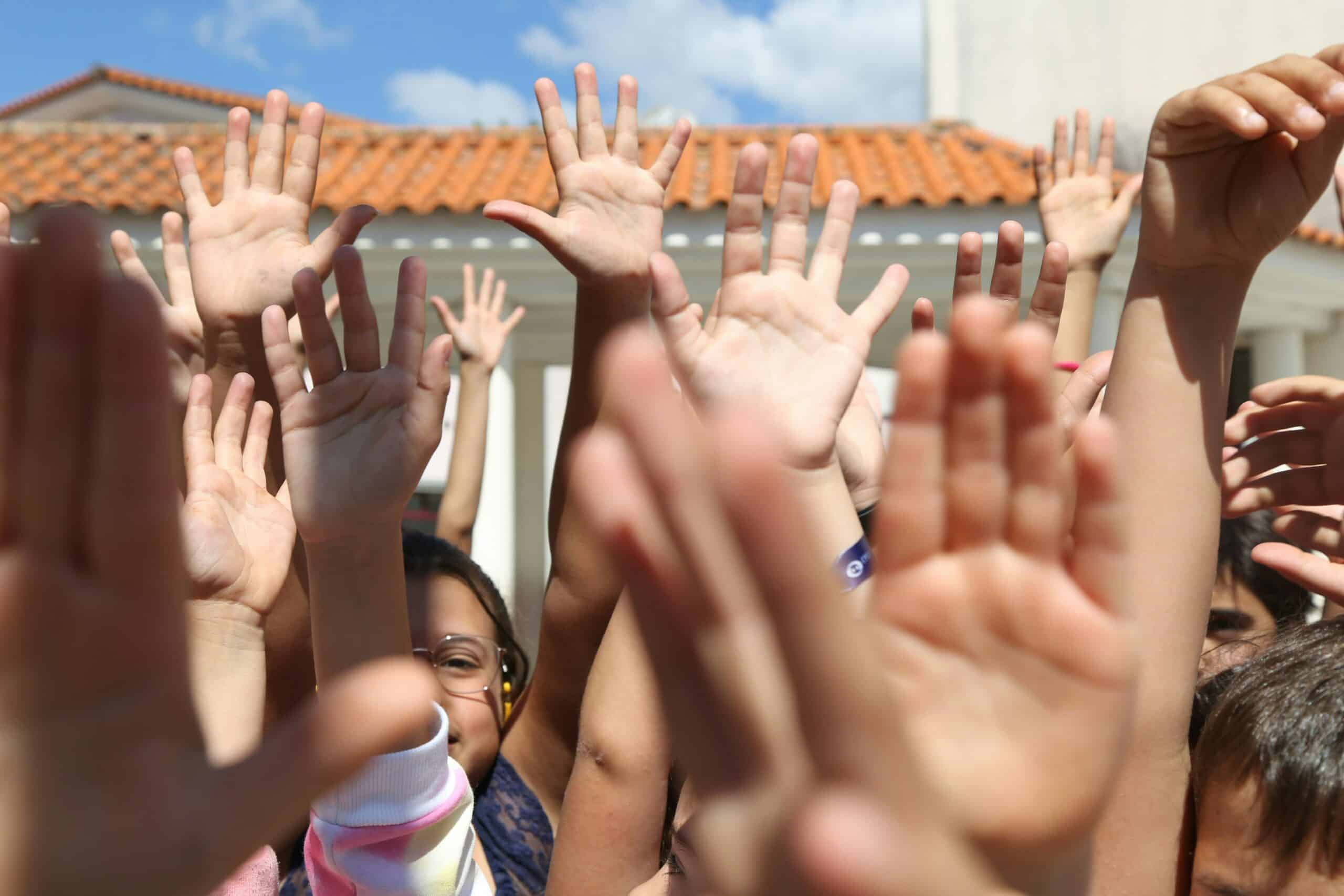 A group of children raise their hands enthusiastically in an outdoor setting, with a bright blue sky and a terracotta-roofed building in the background. Their hands fill the frame, conveying a sense of energy, participation, and unity. Some children’s faces are partially visible, with one wearing glasses and smiling.